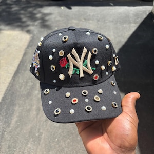 Puede incluir: Una gorra de béisbol negra con un logotipo NY bordado en color canela, una rosa roja con hojas verdes y adornos dispersos que incluyen perlas blancas de imitación y varios diamantes de imitación circulares en engastes dorados, negros y rojos.