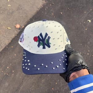 Puede incluir: Gorra de béisbol blanca y azul marino con el logo de los Yankees de Nueva York y una rosa roja. La gorra está adornada con pequeños brillantes. La visera es azul marino.