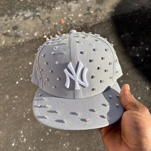 Op de afbeelding: Grijze baseballcap met het witte New York Yankees logo. De cap is versierd met talrijke kleine, glinsterende strass-steentjes. De klep is plat. De cap wordt door een persoon vastgehouden.