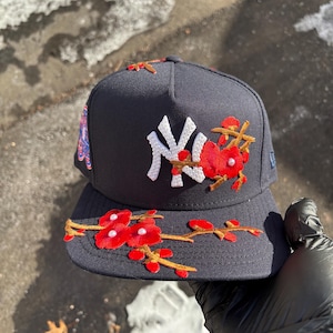 Op de afbeelding: Marineblauwe baseballcap met het New York Yankees-logo in wit. De cap is versierd met geborduurde rode bloemen en takken. Een kleurrijke patch is zichtbaar aan de zijkant.