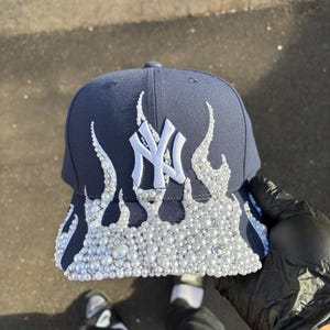 Może przedstawiać: Granatowa czapka z daszkiem z białym logo "NY" i wzorem płomieni. Płomienie zdobione są białymi perłami i dżetami. Klasyczny styl czapki, odpowiedni do noszenia na co dzień.