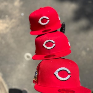 Puede incluir: Tres gorras de béisbol rojas apiladas. Cada gorra presenta un logotipo "C" blanco en la parte delantera. Las gorras están hechas de un material liso y tienen un ajuste estructurado. La gorra inferior tiene un parche en el lateral.
