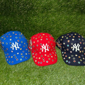Könnte beinhalten: Drei Baseballkappen in Blau, Rot und Marineblau. Jede Kappe hat ein weißes "NY"-Logo und ist mit bunten Strasssteinen verziert. Die Kappen werden auf grünem Gras präsentiert.