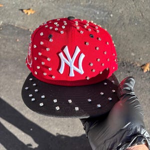 May include: A red baseball cap with a black peak, decorated with multiple square rhinestones. The cap displays the white "NY" logo. The peak and the top of the cap are also embellished with rhinestones.