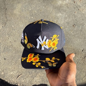 Op de afbeelding: Zwarte baseballcap met het New York Yankees-logo in wit. De cap is versierd met geborduurde gele bloemen en vlinders. De rand en de kroon hebben bloemen- en vlinderontwerpen.