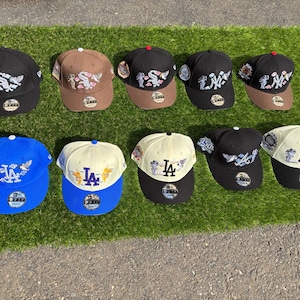 Puede incluir: Una colección de nueve gorras de béisbol dispuestas en dos filas, con varios logotipos de equipos como los New York Yankees, Los Angeles Dodgers y Chicago White Sox. Cada gorra muestra gráficos bordados de querubines y parches laterales.
