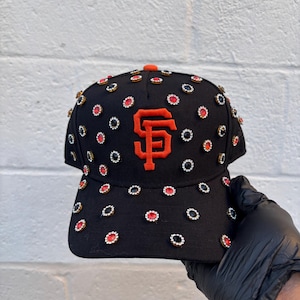 May include: Black baseball cap with the San Francisco Giants logo in orange. The cap is adorned with numerous decorative embellishments in red, white, and black. The brim and top button are orange.