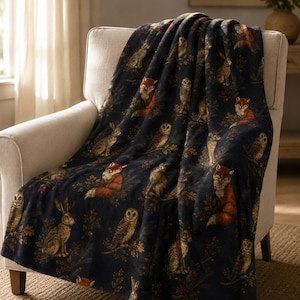 May include: A dark navy blue throw blanket featuring a repeating pattern of orange foxes, brown owls, and rabbits nestled among dark green foliage and small red berries. The blanket is draped over a light beige upholstered armchair.