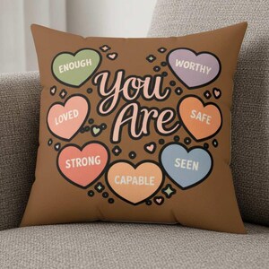 Può includere: Un cuscino quadrato marrone con le parole "You Are" in corsivo, circondato da grafiche a forma di cuore. Ogni cuore contiene un'affermazione positiva: Enough, Worthy, Loved, Safe, Strong, Seen e Capable.