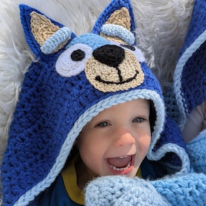 Peut inclure: Un chapeau à capuche et des mitaines assorties en crochet bleu avec un motif de visage de chien. Le chapeau présente de grandes oreilles, un museau beige et un nez noir. L'enfant porte une chemise bleue et sourit.