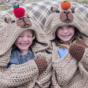 Op de afbeelding: Twee kinderen dragen bijpassende gehaakte berenhoodie-dekens. De dekens zijn bruin met bruine berengezichten op de capuchons, compleet met kleine, gehaakte appels en sinaasappels erop. De kinderen lachen, gehuld in de gezellige dekens.