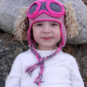 May include: A pink and gray crocheted hat with goggles and ear flaps. The hat has a braided strap that ties under the chin. The hat is worn by a child.