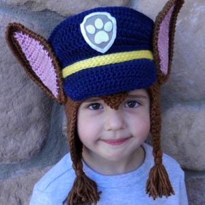 May include: A blue and brown crocheted hat with a paw print badge and yellow stripe. The hat has long brown ear flaps and tassels.