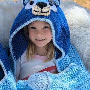 K&ouml;nnte beinhalten: Eine blaue H&auml;keldecke mit Kapuze und einem Hundegesicht-Design. Die Decke hat eine Kapuze mit Ohren, Augen und einer Schnauze. Die Decke ist eine Mischung aus hell- und dunkelblauem Garn mit hellblauer Bord&uuml;re.