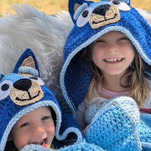 K&ouml;nnte beinhalten: Zwei Kinder tragen handgefertigte blaue H&auml;keldecken mit Hundefratzen. Die Decken haben hellblaue und beige Akzente. Die Kinder l&auml;cheln, der Hintergrund ist unscharf.