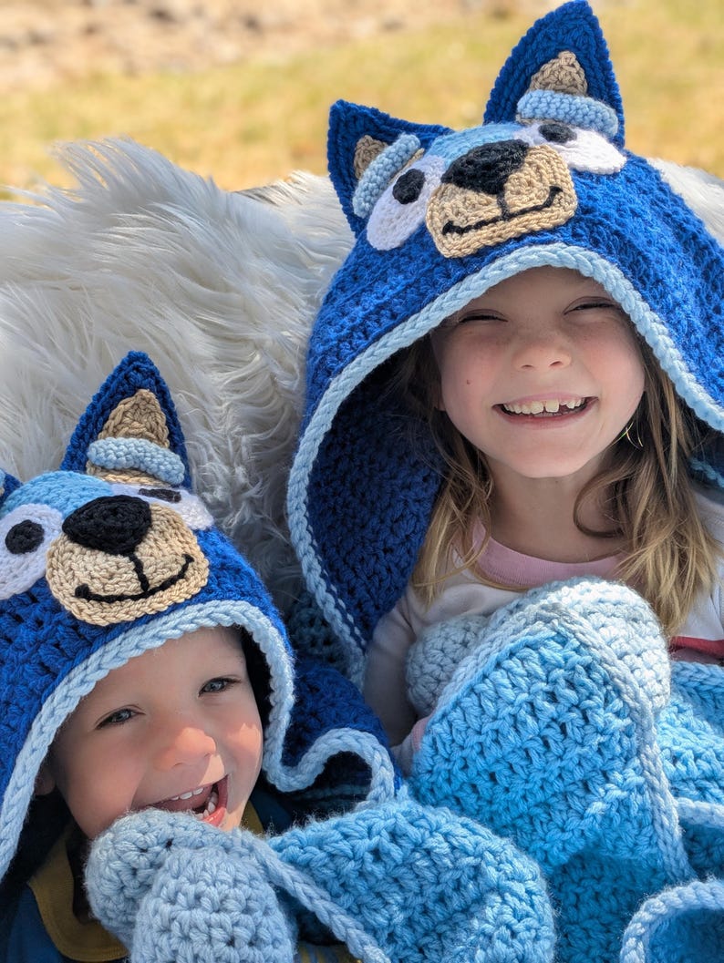 K&ouml;nnte beinhalten: Zwei Kinder tragen handgefertigte blaue H&auml;keldecken mit Hundefratzen. Die Decken haben hellblaue und beige Akzente. Die Kinder l&auml;cheln, der Hintergrund ist unscharf.