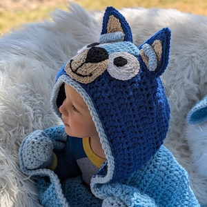 K&ouml;nnte beinhalten: Ein blaues geh&auml;keltes Kapuzen-Decken- und M&uuml;tzen-Set. Die M&uuml;tze zeigt ein Cartoon-Hunde-Gesicht mit braunen, schwarzen und wei&szlig;en Details. Die Decke und die M&uuml;tze sind in Blaut&ouml;nen gehalten. Das Set ist f&uuml;r ein Kind konzipiert.