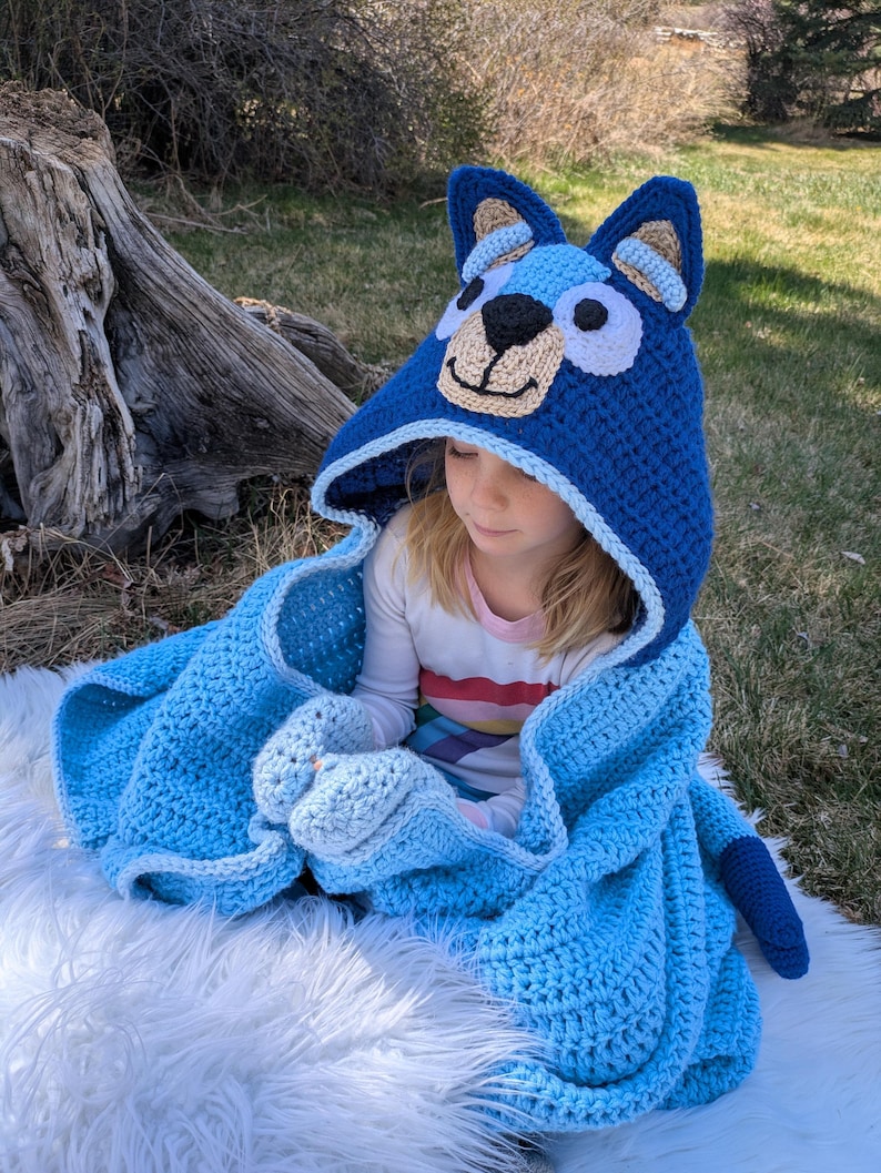 K&ouml;nnte beinhalten: Eine blaue H&auml;keldecke mit Kapuze und einem Hundegesicht-Design. Die Kapuze hat eine hellbraune Schnauze, eine schwarze Nase und blaue Ohren. Die Decke ist in einem helleren Blauton gehalten und hat passende F&auml;ustlinge.