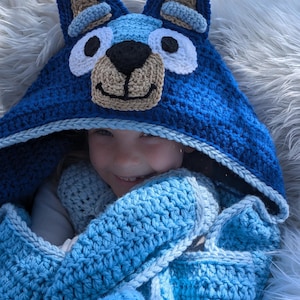 K&ouml;nnte beinhalten: Eine handgefertigte blaue H&auml;keldecke mit Kapuze und einem Hundegesicht-Design. Die Kapuze hat gro&szlig;e Ohren, wei&szlig;e Augen, eine schwarze Nase und eine beige Schnauze. Die Decke hat einen Farbverlauf von hell- zu dunkelblau, ideal f&uuml;r Kinder.