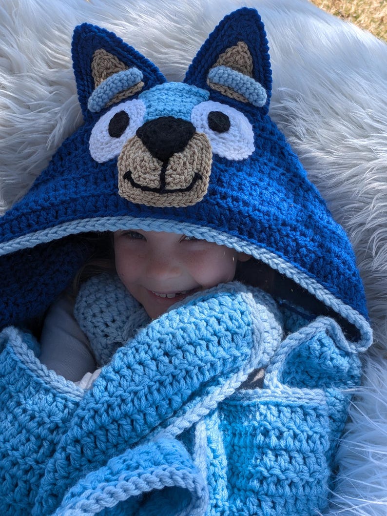 K&ouml;nnte beinhalten: Eine handgefertigte blaue H&auml;keldecke mit Kapuze und einem Hundegesicht-Design. Die Kapuze hat gro&szlig;e Ohren, wei&szlig;e Augen, eine schwarze Nase und eine beige Schnauze. Die Decke hat einen Farbverlauf von hell- zu dunkelblau, ideal f&uuml;r Kinder.