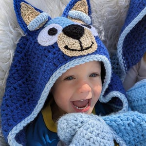 K&ouml;nnte beinhalten: Ein blauer geh&auml;kelter Kapuzenhut und passende F&auml;ustlinge mit einem Cartoon-Hunde-Gesicht. Der Hut hat gro&szlig;e Ohren, eine beige Schnauze und eine schwarze Nase. Die F&auml;ustlinge und der Hut sind in Blau- und Wei&szlig;t&ouml;nen gehalten.