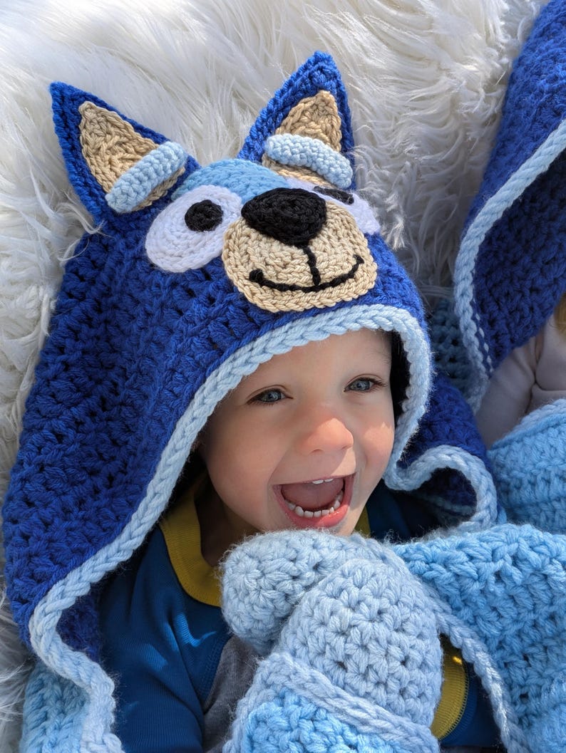 K&ouml;nnte beinhalten: Ein blauer geh&auml;kelter Kapuzenhut und passende F&auml;ustlinge mit einem Cartoon-Hunde-Gesicht. Der Hut hat gro&szlig;e Ohren, eine beige Schnauze und eine schwarze Nase. Die F&auml;ustlinge und der Hut sind in Blau- und Wei&szlig;t&ouml;nen gehalten.