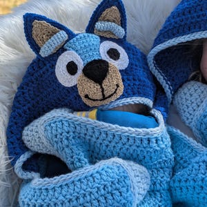 K&ouml;nnte beinhalten: Eine handgefertigte blaue H&auml;keldecke mit Kapuze und einem Hundegesicht-Design. Die Kapuze hat gro&szlig;e Ohren, wei&szlig;e Augen und eine hellbraune Schnauze. Die Decke ist eine Mischung aus hell- und dunkelblauem Garn und sorgt f&uuml;r einen gem&uuml;tlichen und verspielten Look.