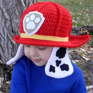 Könnte beinhalten: Ein roter gehäkelter Hut mit gelbem Band und einem weißen und grauen Pfotenabdruck-Emblem. Der Hut hat weiße Ohrenklappen mit schwarzen Flecken. Der Hut ist als Feuerwehrhut gestaltet.