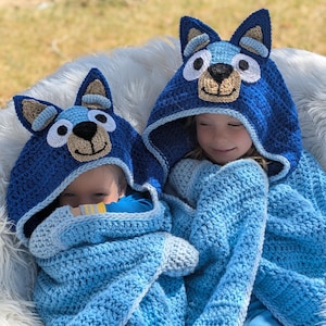 K&ouml;nnte beinhalten: Zwei Kinder tragen blaue geh&auml;kelte Kapuzen-Decken mit hundeartigen Ohren und Gesichtern. Die Decken sind in Blau- und Hellblaut&ouml;nen gehalten. Die Kinder sitzen auf einer wei&szlig;en, pelzigen Oberfl&auml;che mit verschwommenem gr&uuml;nem Hintergrund.