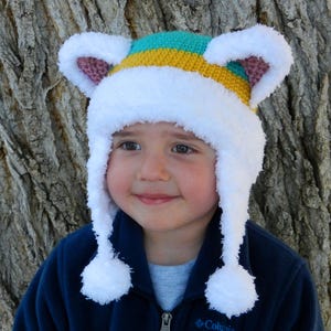 May include: A white crocheted hat with fuzzy ear flaps and a striped brim in teal, yellow, and white. The hat has white pom-poms on the ends of the ear flaps.