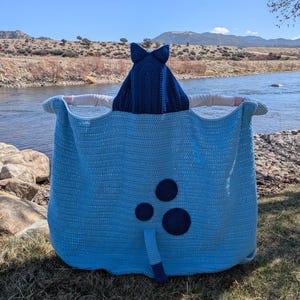 K&ouml;nnte beinhalten: Eine hellblaue H&auml;keldecke mit einer dunkelblauen Kapuze und Akzenten. Die Decke hat einen Schwanz und drei dunkelblaue Kreise. Die Kapuze hat zwei spitze Ohren. Die Decke befindet sich im Freien in der N&auml;he eines Flusses.