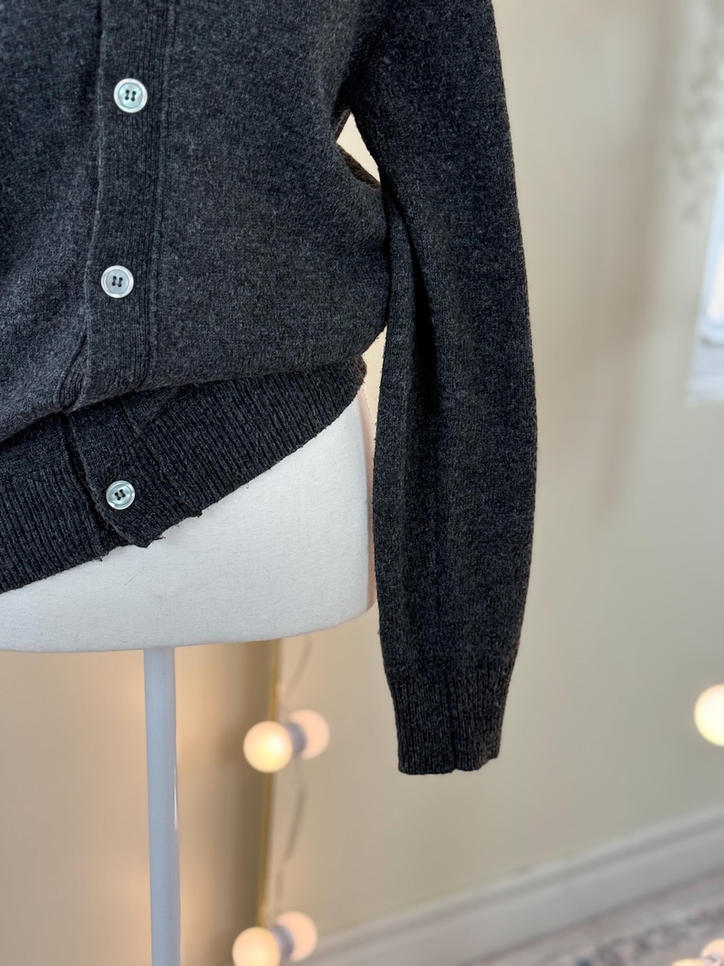 May include: Dark gray cardigan sweater with a ribbed hem and cuffs. The sweater has a button-up front with three white buttons. The sleeve is long and the fabric appears to be a soft knit.