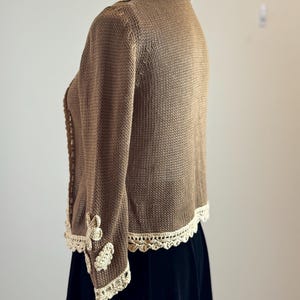 May include: A brown knitted cardigan with cream-colored crochet trim along the collar, cuffs, and hem. The sleeves feature cream-colored floral appliqu&eacute;s. The cardigan is displayed on a mannequin, paired with a black skirt.