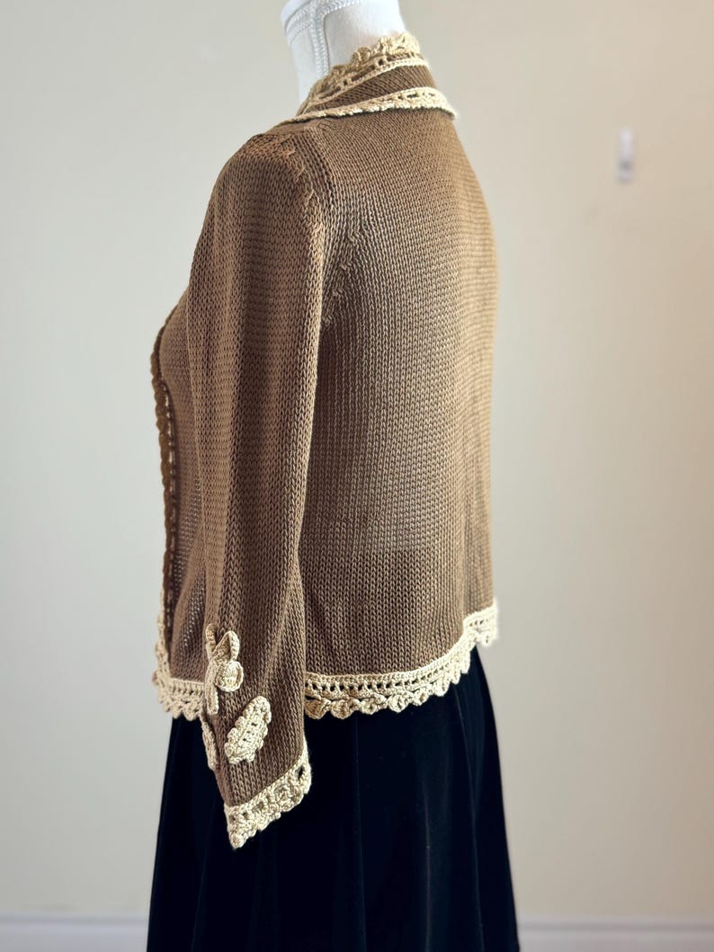 May include: A brown knitted cardigan with cream-colored crochet trim along the collar, cuffs, and hem. The sleeves feature cream-colored floral appliqu&eacute;s. The cardigan is displayed on a mannequin, paired with a black skirt.