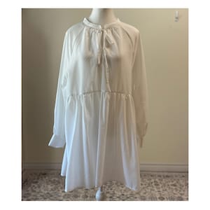 Puede incluir: Un vestido blanco de manga larga con cuello alto y detalle de lazo. El vestido presenta una cintura fruncida y una falda acampanada. Las mangas tienen puños abotonados. El vestido se exhibe en un maniquí.