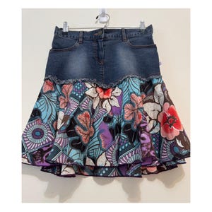 Peut inclure: Jupe en jean avec imprimé floral. La jupe présente une ceinture en jean bleu foncé et une jupe évasée avec un motif floral coloré dans les tons de bleu, violet, rose et blanc. L'ourlet du jean est effiloché.
