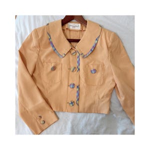 Può includere: Giacca color pesca con colletto arrotondato e due tasche frontali. Il colletto e l'abbottonatura sono rifiniti con un motivo a quadri colorati. La giacca ha tre bottoni su ogni manica e un'etichetta che dice "Mary Jane".