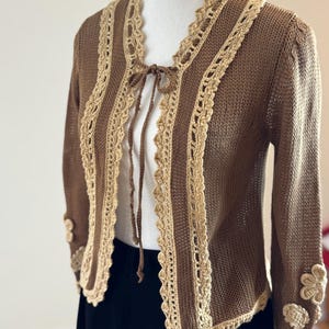 May include: A brown knit cardigan with a cream-colored crochet trim and tie closure. The long-sleeved sweater features floral embellishments on the cuffs. The open front design is accented with a cream-colored crochet trim.