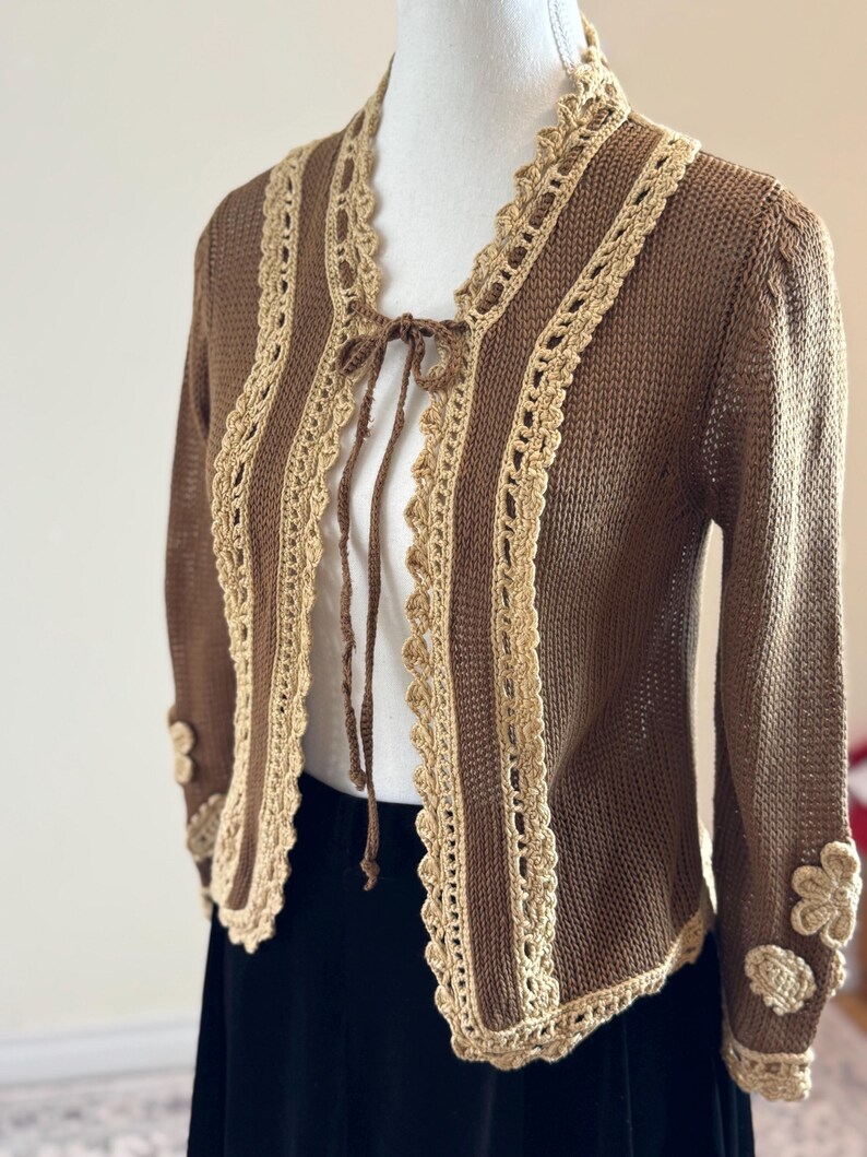 May include: A brown knit cardigan with a cream-colored crochet trim and tie closure. The long-sleeved sweater features floral embellishments on the cuffs. The open front design is accented with a cream-colored crochet trim.