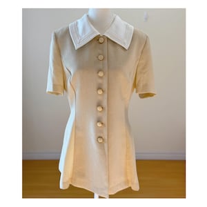 May include: A beige short-sleeved blouse with a white collar. The collar has a decorative trim. The blouse has a button-up front with six beige buttons. The garment is displayed on a mannequin.