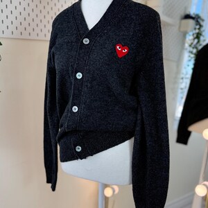 May include: Dark gray cardigan sweater with a V-neck and button closure. The sweater features a red heart-shaped patch with eyes on the left chest. The sweater is displayed on a mannequin.