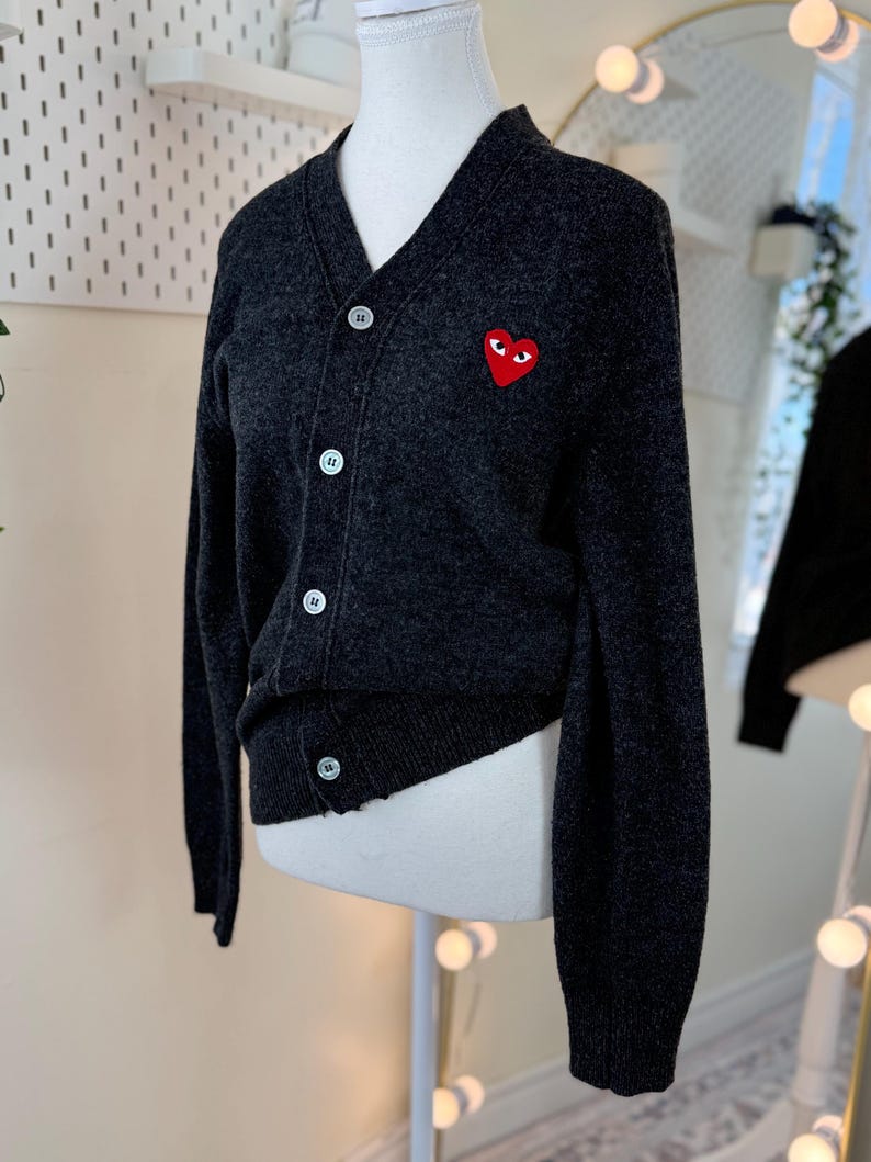 May include: Dark gray cardigan sweater with a V-neck and button closure. The sweater features a red heart-shaped patch with eyes on the left chest. The sweater is displayed on a mannequin.