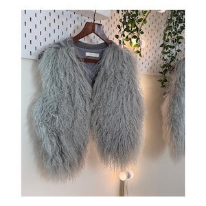 May include: A light grey, open-front vest with a shaggy, textured appearance. The vest is hanging on a wooden hanger. The vest appears to be made of a soft, possibly faux fur material. The background is a white wall.
