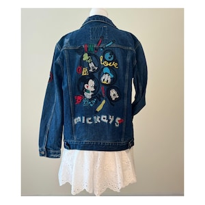 May include: A dark blue denim jacket with colorful embroidered patches featuring Mickey Mouse and friends. The back of the jacket has the words "IN LOVE OR MICKEYS" in a playful font. The jacket has a classic button-up design.