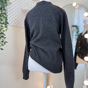 May include: A dark gray sweater displayed on a white mannequin. The sweater has long sleeves and a crew neck. The fabric appears to be a soft knit. The mannequin is positioned in front of a white pegboard and a mirror.