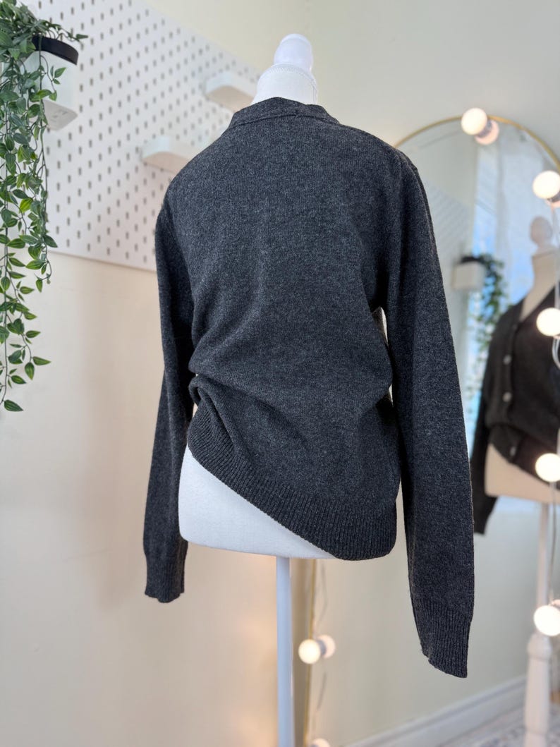 May include: A dark gray sweater displayed on a white mannequin. The sweater has long sleeves and a crew neck. The fabric appears to be a soft knit. The mannequin is positioned in front of a white pegboard and a mirror.