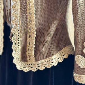 May include: A tan knitted cardigan with cream-colored crochet trim. The trim features a scalloped edge and is visible along the front and sleeve cuffs. The cardigan is made of a soft, textured material.