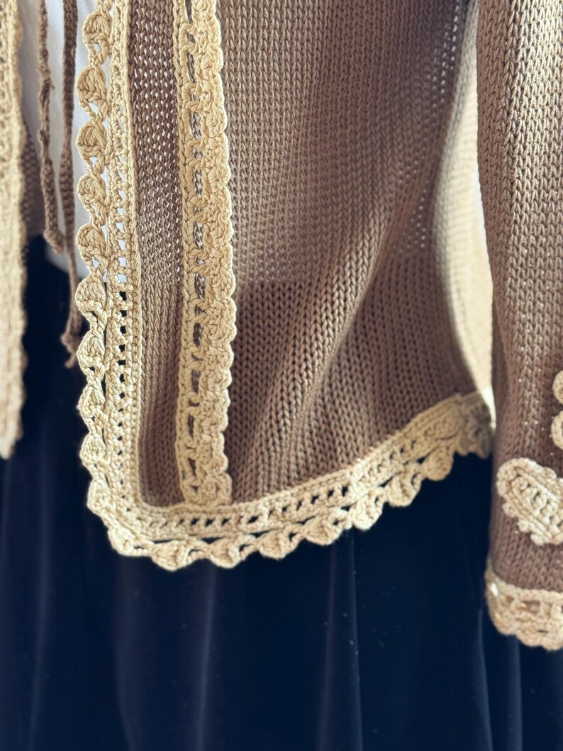 May include: A tan knitted cardigan with cream-colored crochet trim. The trim features a scalloped edge and is visible along the front and sleeve cuffs. The cardigan is made of a soft, textured material.