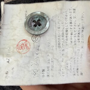 May include: Close-up of a garment label with Japanese text, a large button, and a red stamp. The button is a light gray color with four holes. The label has the number 25 and the text "AZ T184".