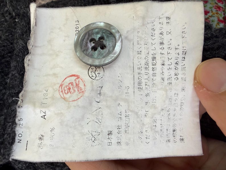 May include: Close-up of a garment label with Japanese text, a large button, and a red stamp. The button is a light gray color with four holes. The label has the number 25 and the text "AZ T184".